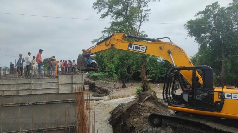 JCB के पंजे पर बैठकर नदी का पुल पार कर रहे लोग
